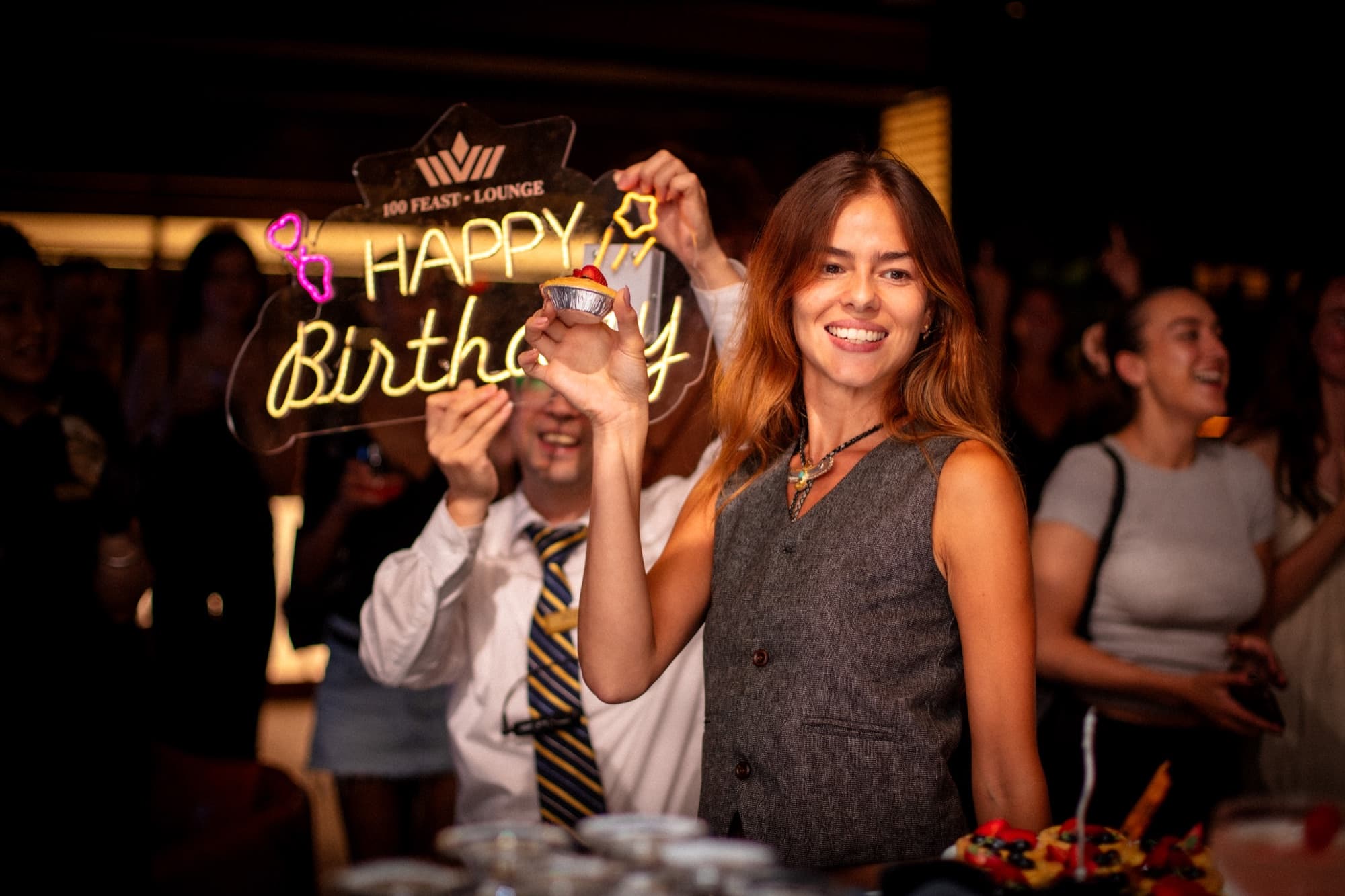 A guest holding a lit birthday tart beneath the lounge's neon Happy Birthday sign